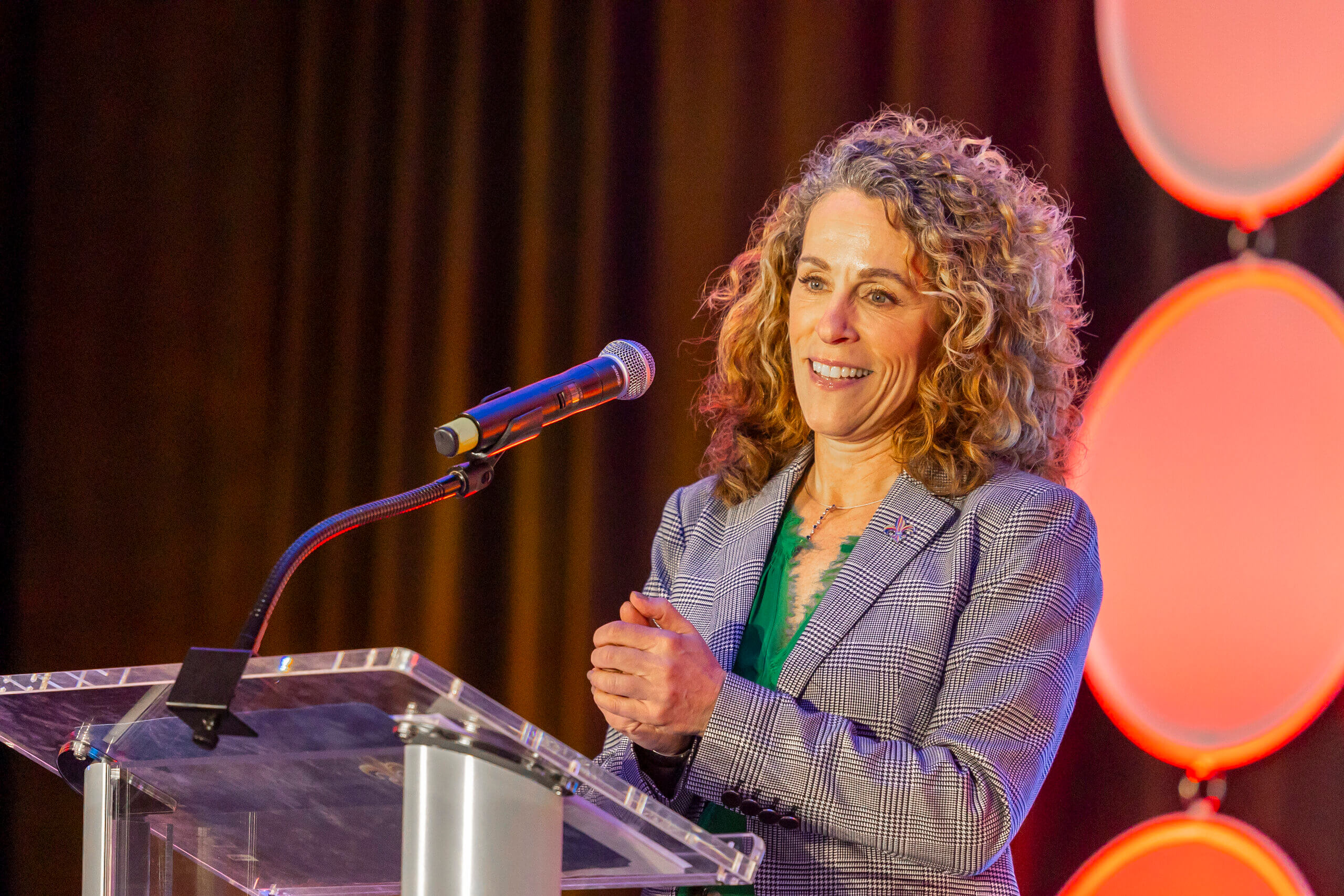 A woman with curly hair, stands at a podium speaking into a microphone, framed by circular red and white lights in the background.