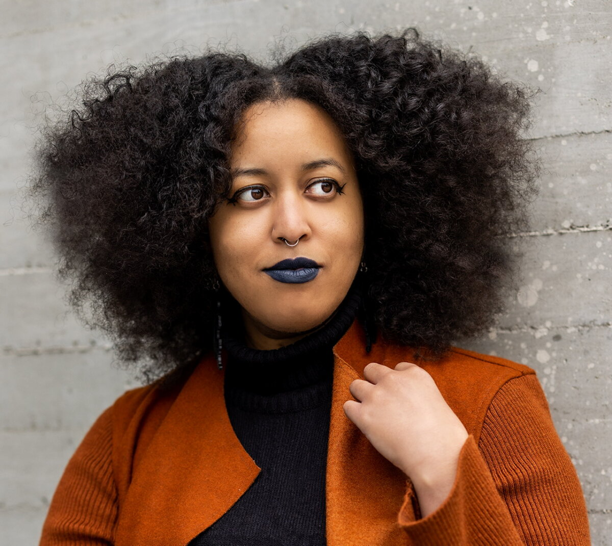 How Louisville Artist Robyn Gibson Turns Boxing Into Art - 2 Robyn Gibson stands with natural curly hair, dark lipstick, and a rust-colored coat over a black turtleneck in front of a textured concrete wall.