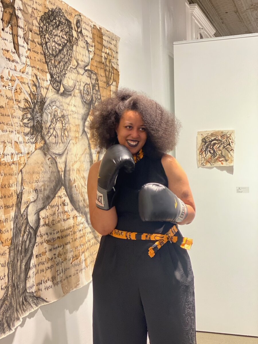 How Louisville Artist Robyn Gibson Turns Boxing Into Art - 4 Robyn Gibson, featured in FACES of Louisville, stands with curly hair and boxing gloves in front of large figurative artwork on a gallery wall, smiling at the camera.