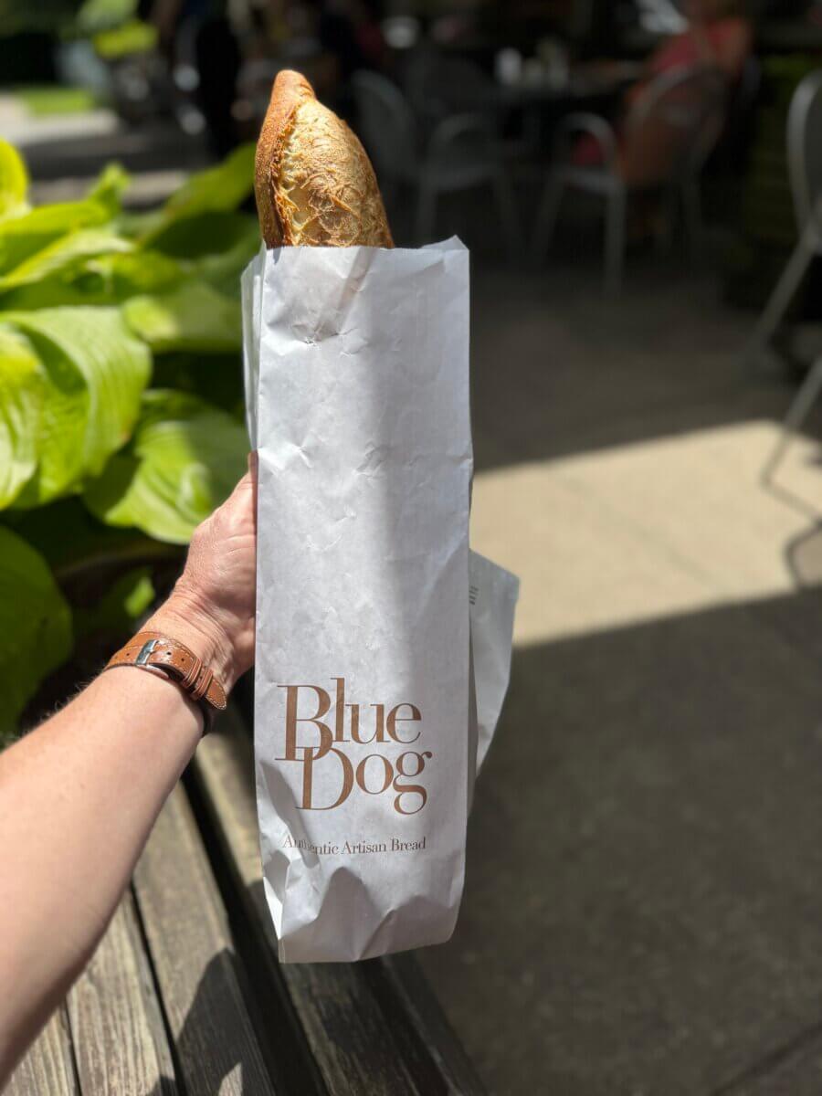 16 Louisville FINDS for June: Summer Style, Tabletop Charm & Little Luxuries - 13 A person holds a baguette in a paper bag labeled "Blue Dog" outdoors in Louisville, with green plants and people enjoying their everyday luxuries at nearby tables in the background.