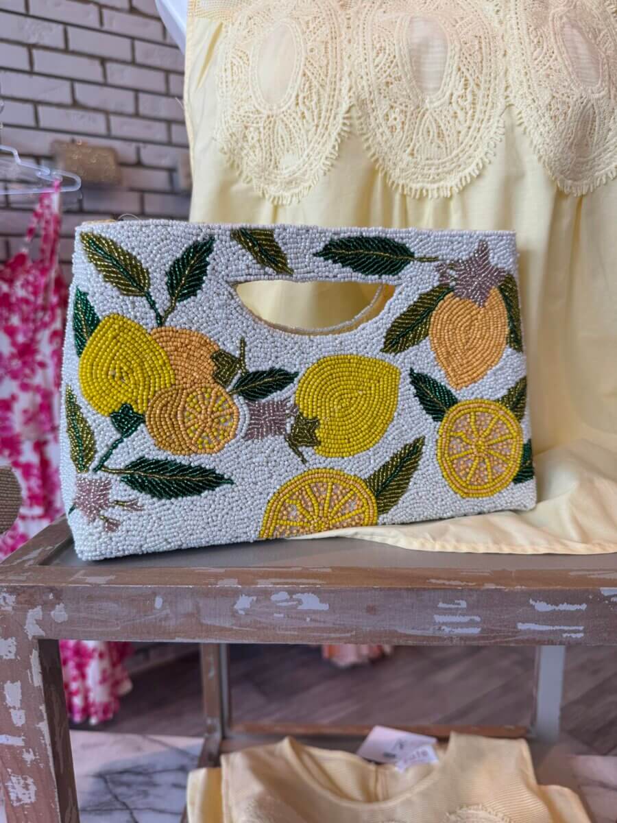16 Louisville FINDS for June: Summer Style, Tabletop Charm & Little Luxuries - 4 A beaded clutch bag featuring yellow lemons and green leaves sits on a wooden table next to light yellow clothing—one of Louisville’s standout summer picks for those who appreciate everyday luxuries.