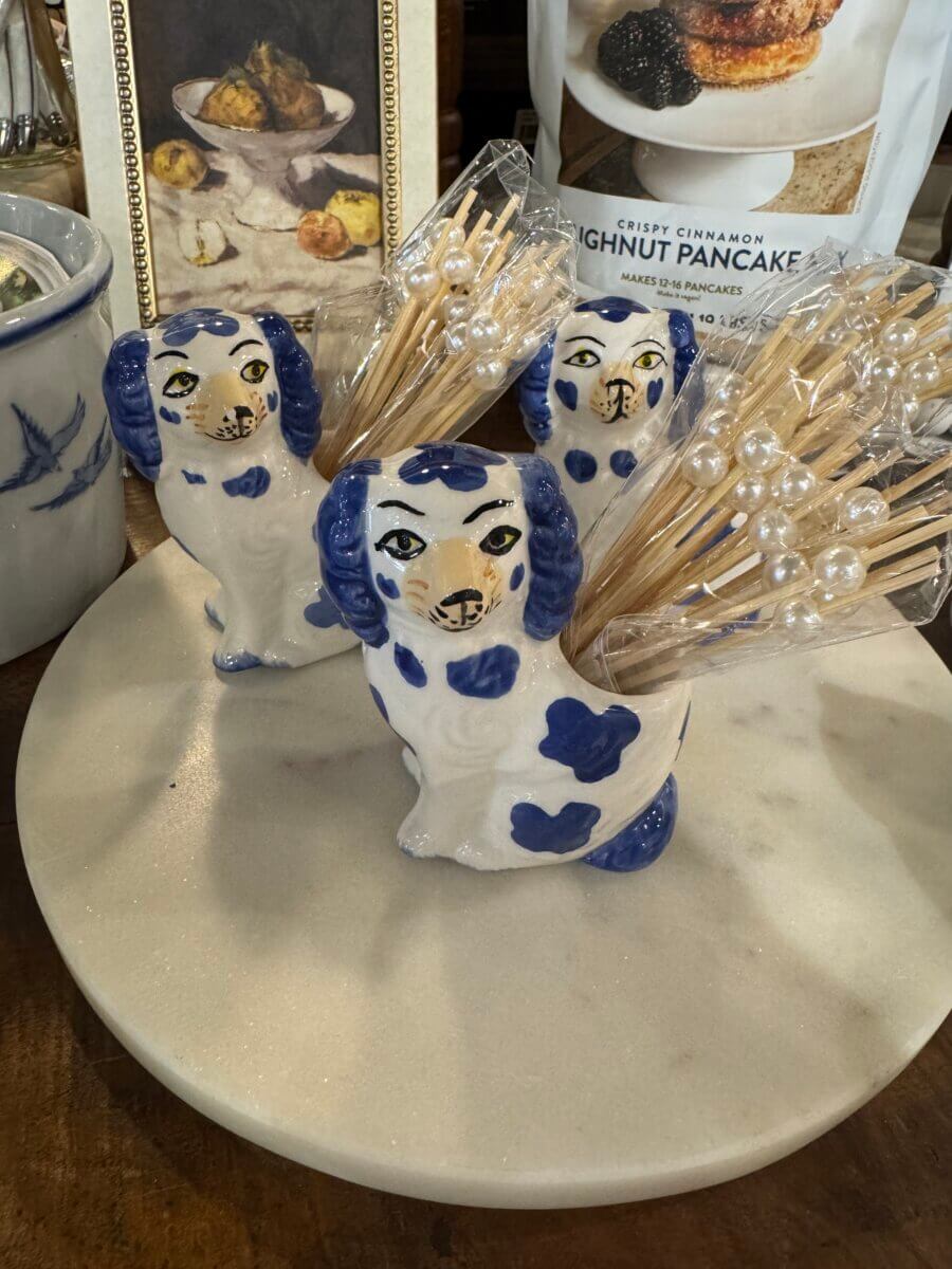 16 Louisville FINDS for June: Summer Style, Tabletop Charm & Little Luxuries - 10 Three ceramic dog figurines with blue spots hold pearl-tipped cocktail picks—perfect for summer picks or everyday luxuries—on a round white tray, with pantry items and framed art in the background.