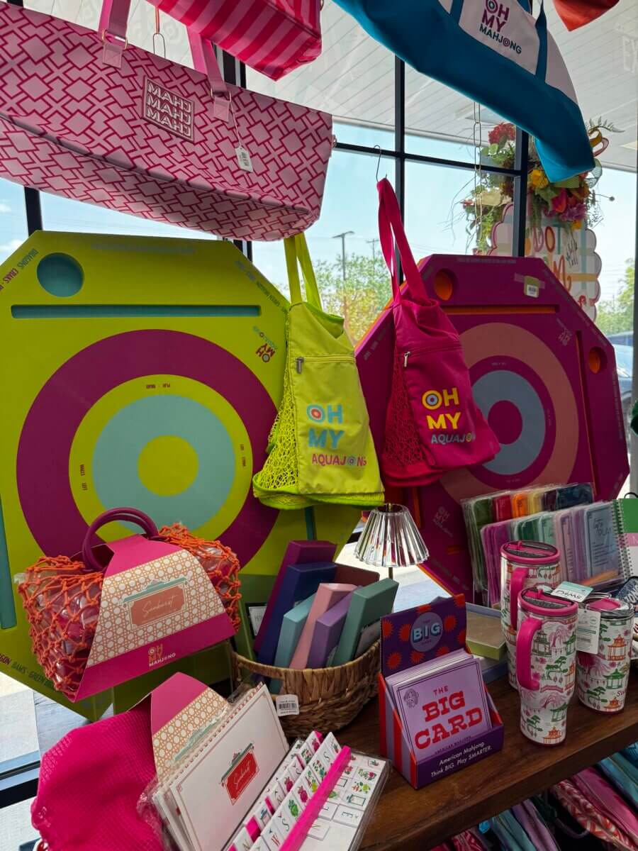 16 Luxe Louisville FINDS for July: Fine Art, Rare Bourbon & Resort-Ready Style - 17 Display of colorful tote bags, round pool floats, stationery, and drinkware arranged on shelves in a brightly lit Luxe Louisville store with large windows.