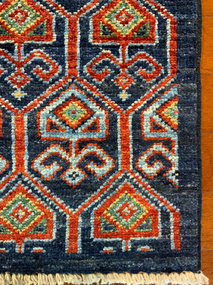 16 Luxe Louisville FINDS for July: Fine Art, Rare Bourbon & Resort-Ready Style - 3 A section of a woven rug with a geometric diamond and floral pattern in red, blue, white, and green on a dark blue background. Featured in Luxe Louisville’s July Louisville FINDS, the rug has fringed ends.