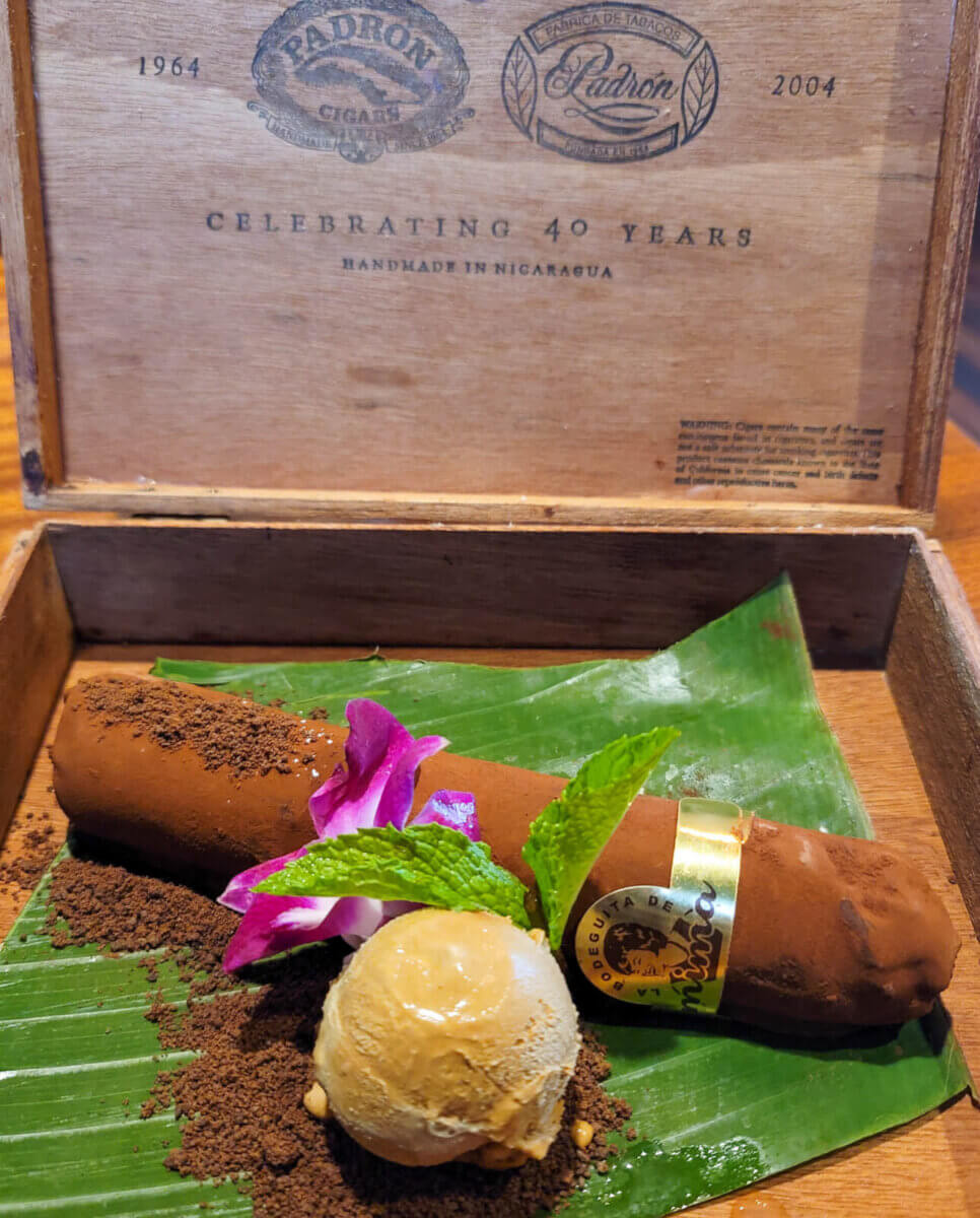 25+ Louisville Restaurants Open on July 4th - 5 A chocolate dessert shaped like a cigar, garnished with cocoa powder, a mint sprig, a purple flower, and caramel ice cream, served on a banana leaf in a wooden cigar box—just one of the creative treats found at top Louisville restaurants.