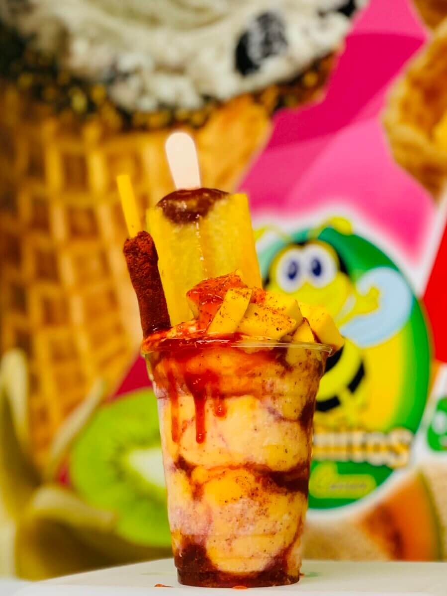 10 Louisville Ice Cream Spots to Beat the Heat - 10 A clear plastic cup filled with a frozen fruit dessert topped with chili powder, chamoy, a tamarind stick, and a yellow popsicle—perfect for summer and inspired by the vibrant treats at top Louisville ice cream spots.