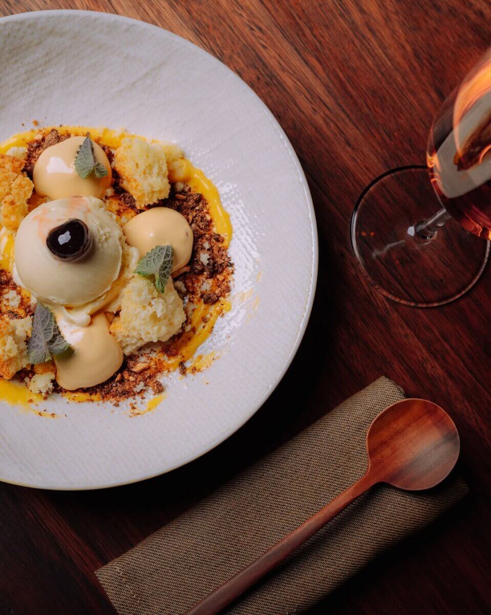 10 Louisville Ice Cream Spots to Beat the Heat - 11 A plated dessert with ice cream, cake pieces, crumbles, sauce, and mint leaves on a white dish, next to a glass of rosé wine and a wooden spoon on a napkin.