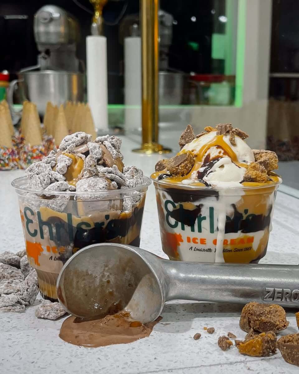 10 Louisville Ice Cream Spots to Beat the Heat - 5 Two cups of Louisville ice cream with chocolate sauce and toppings sit on a counter next to an ice cream scoop and cone ingredients—capturing the sweet flavors of summer.