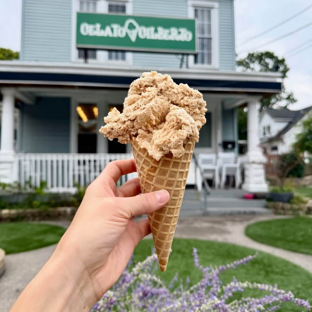 10 Louisville Ice Cream Spots to Beat the Heat - 6 A hand holds a waffle cone with a scoop of light brown gelato in front of a blue building with a "Gelato" sign—one of the must-visit Louisville ice cream spots for summer treats.