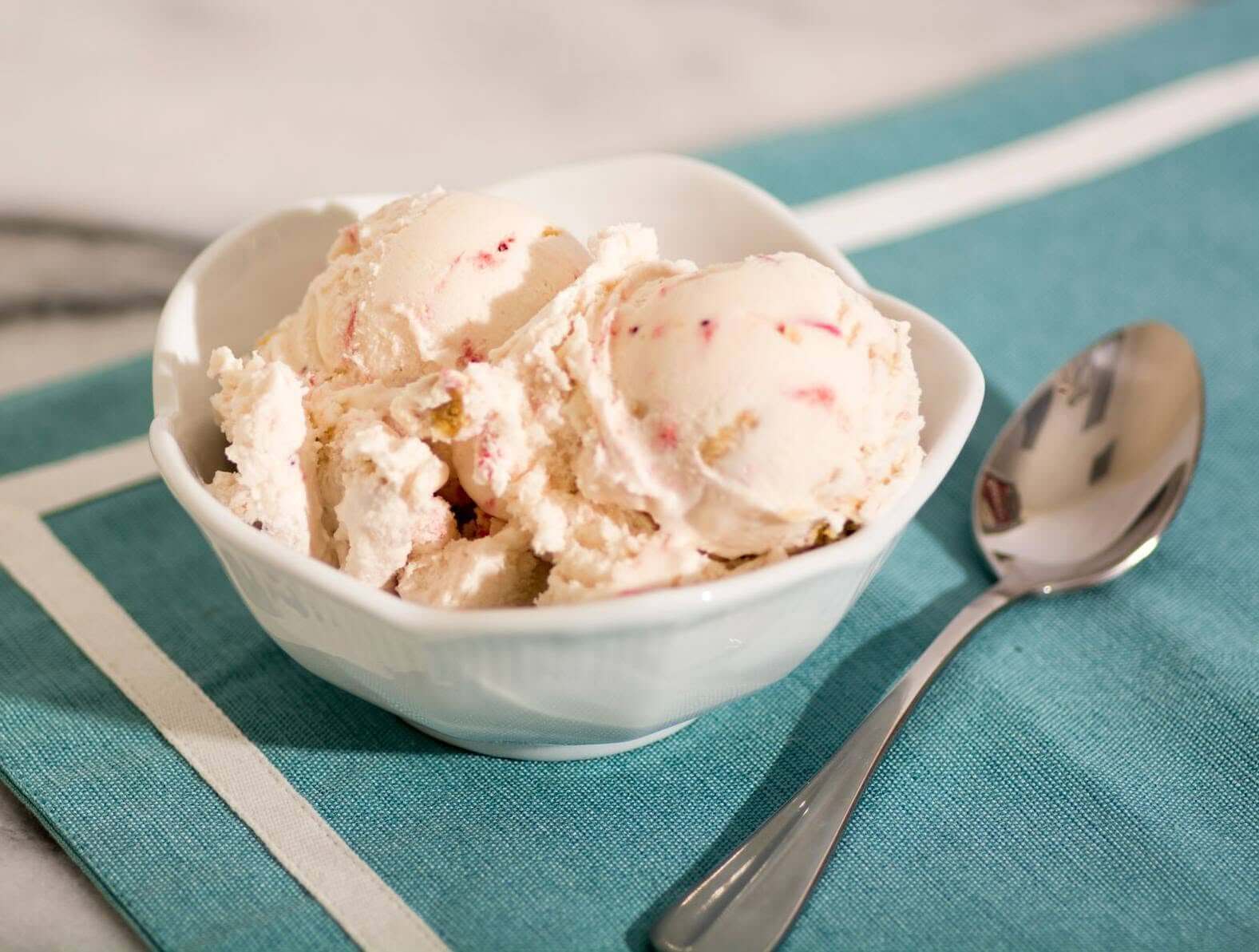 A white bowl with two scoops of ice cream, possibly with fruit pieces, placed on a blue napkin next to a metal spoon.