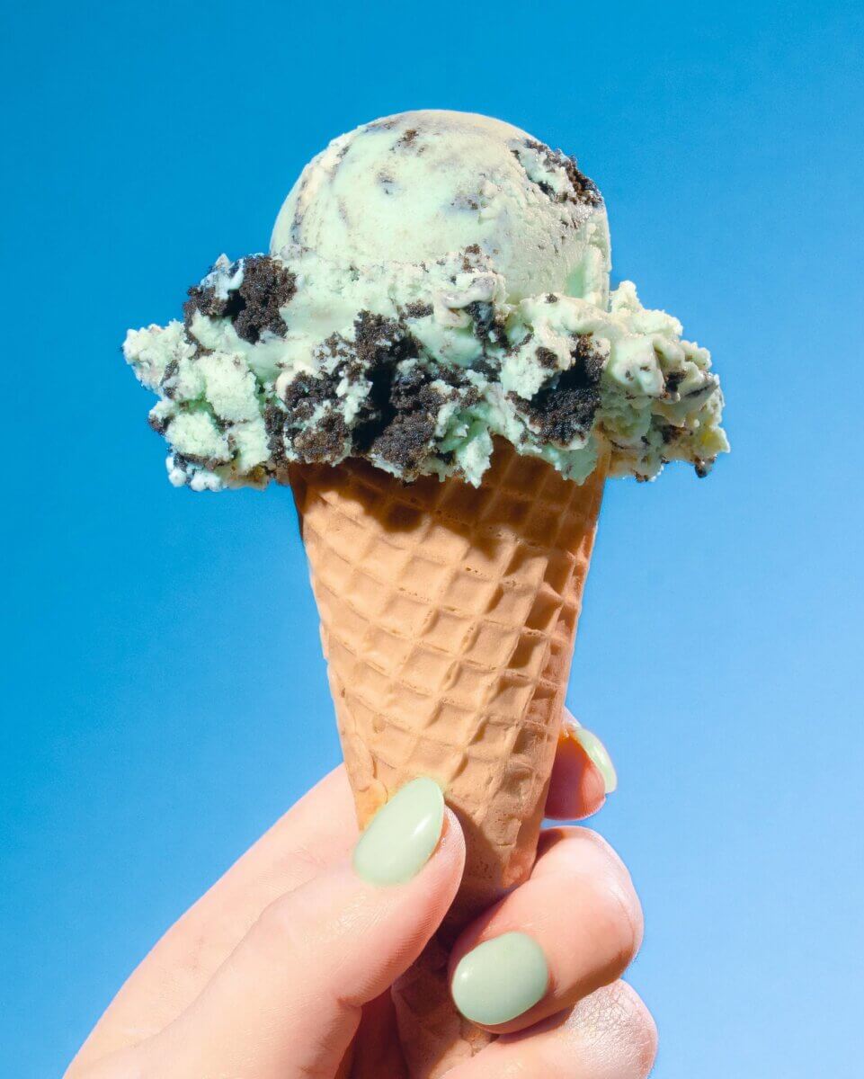 10 Louisville Ice Cream Spots to Beat the Heat - 7 A hand with mint green nail polish holds a waffle cone with a scoop of mint chocolate chip ice cream against a blue background, capturing the perfect summer vibe.