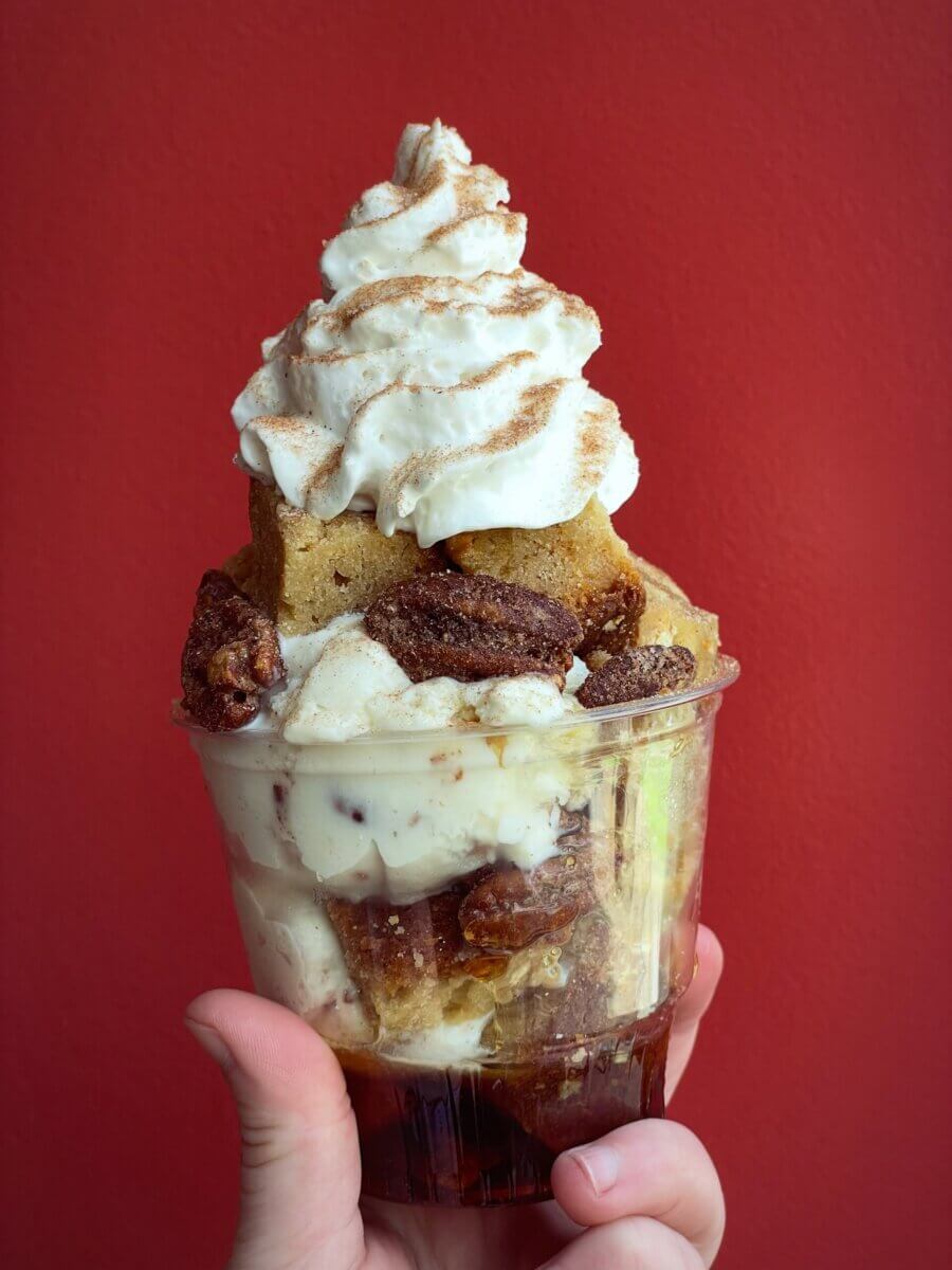 10 Louisville Ice Cream Spots to Beat the Heat - 9 A hand holds a clear cup filled with Louisville ice cream, cake pieces, candied pecans, whipped cream, and cinnamon against a plain red background.