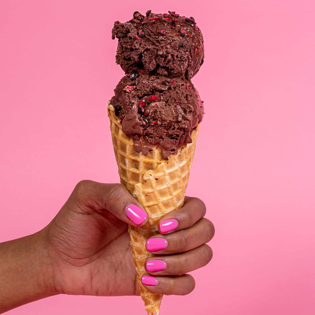 10 Louisville Ice Cream Spots to Beat the Heat - 2 A hand with pink nail polish holds a waffle cone with two scoops of chocolate ice cream against a pink background, capturing the sweet taste of summer and the joy of discovering new Louisville ice cream spots.