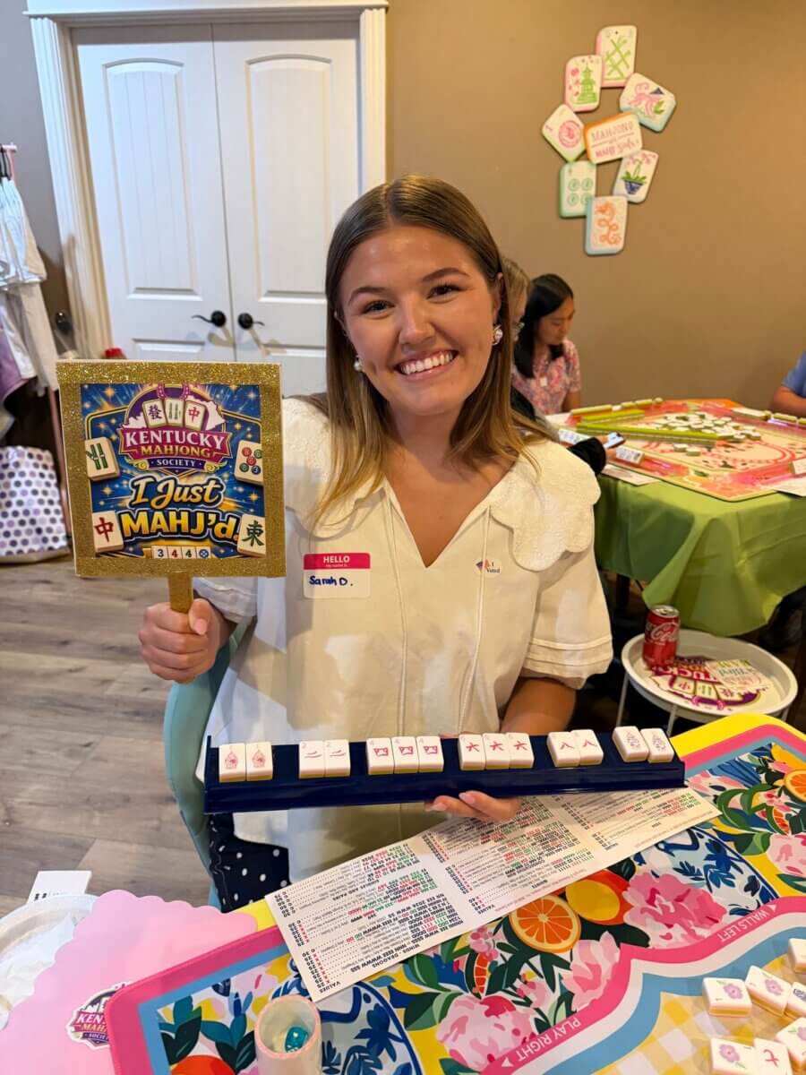 Your Ultimate Louisville Mahjong Guide - 3 A woman smiles while holding a βKentucky I just MAHJ'd!β sign and displaying mahjong tiles at a decorated game table in Louisville.