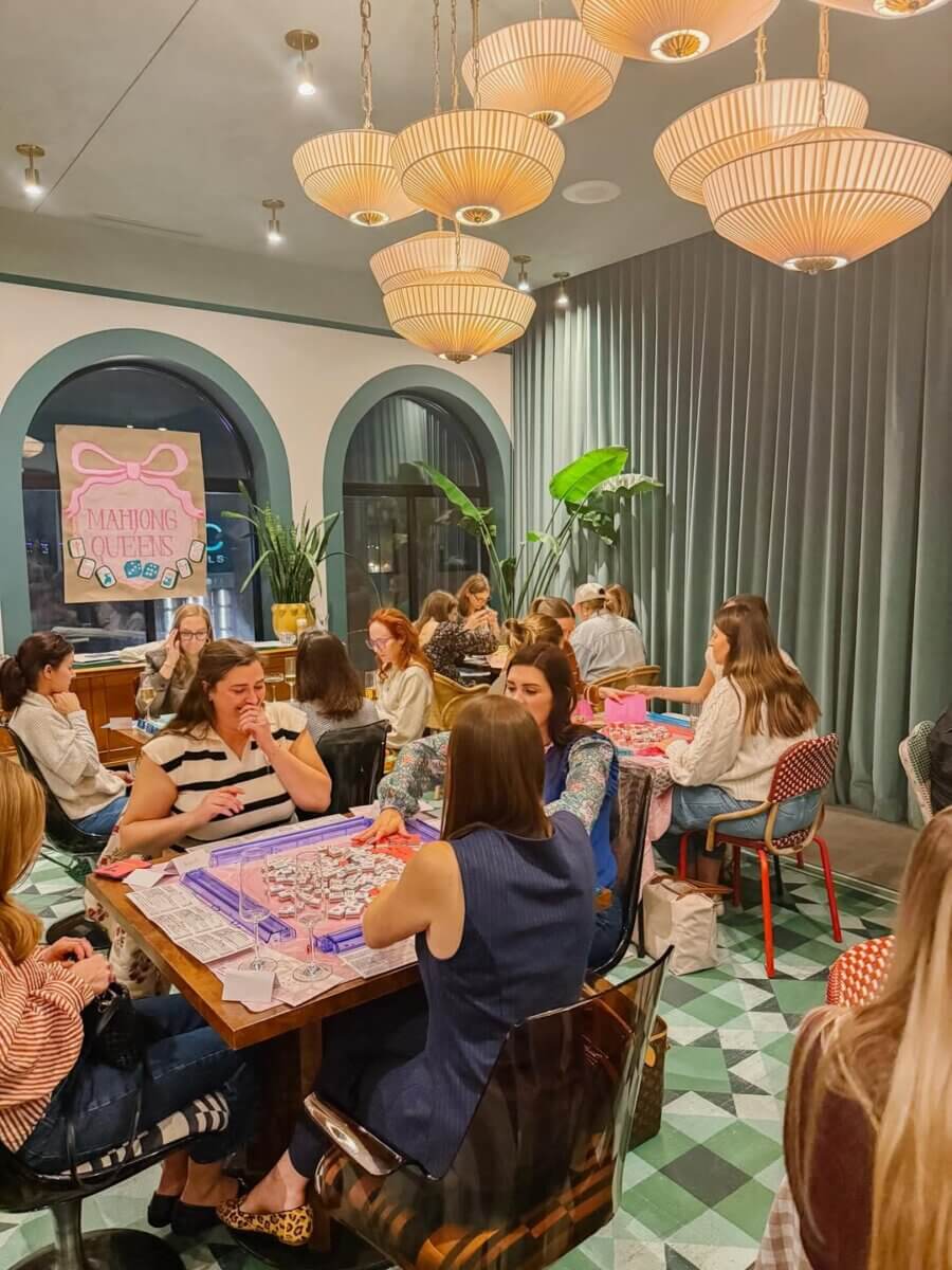 Your Ultimate Louisville Mahjong Guide - 2 A group of women sit at tables playing Mahjong in a well-lit, modern Louisville room with green curtains and large pendant lights.