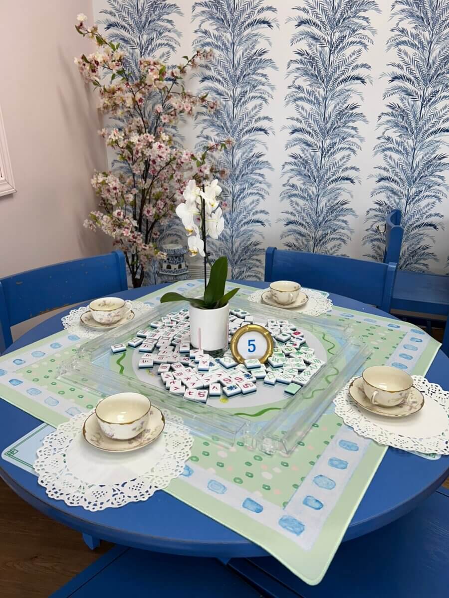 Your Ultimate Louisville Mahjong Guide - 4 A blue round table set for four with teacups, saucers, doilies, a Mahjong set in the center, an orchid plant, and a "5" table number; floral wallpaper and a blossoming plant in the background adds Louisville charm.