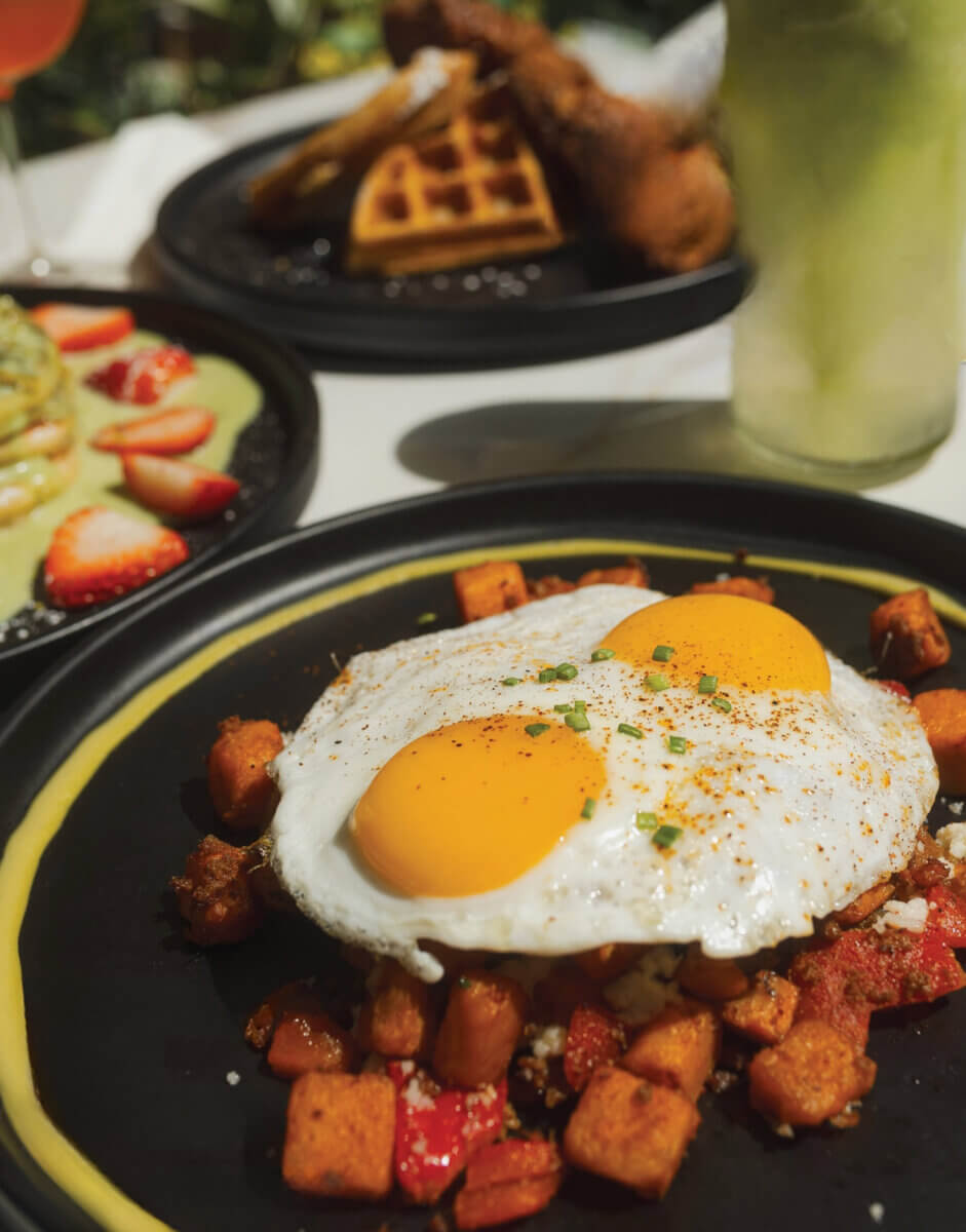 3 New Louisville Restaurants to Try (+2 New Menus!) - 5 A plate with two sunny-side-up eggs served over diced potatoes, with two additional plates of food and a green drink in the background, showcases the delicious offerings at new Louisville restaurants.