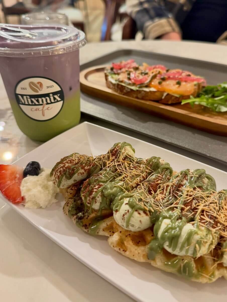 3 New Louisville Restaurants to Try (+2 New Menus!) - 2 A plate of savory waffles topped with green sauce and dried garnish, a side of fruit and cream, a gradient drink in a Mixmix Cafe cup, and an open-faced sandwich in the background.