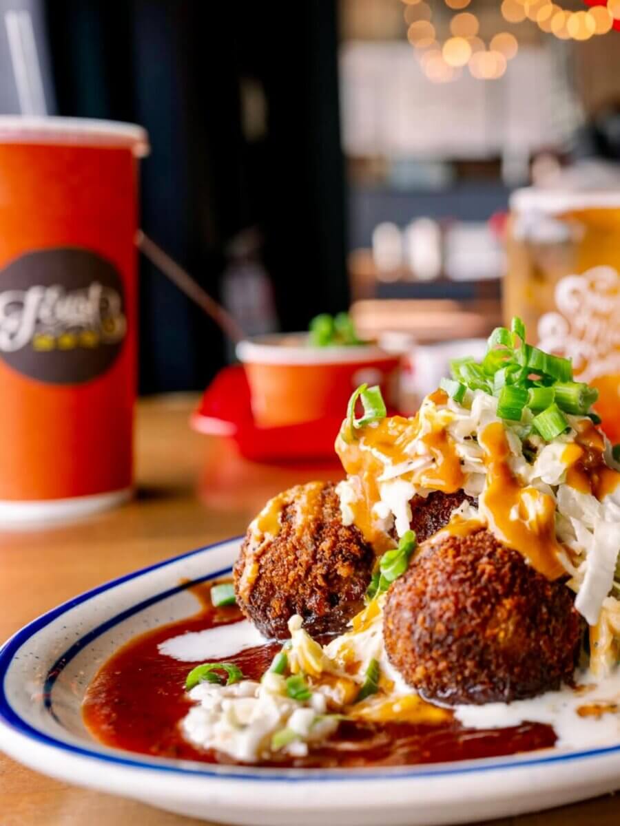 25+ Louisville Restaurants Open on July 4th - 4 Three fried croquettes topped with coleslaw, chopped green onions, and sauce, served on a plate with brown gravy—classic comfort from Louisville restaurants, perfect for enjoying this July 4th. A drink cup is in the background.