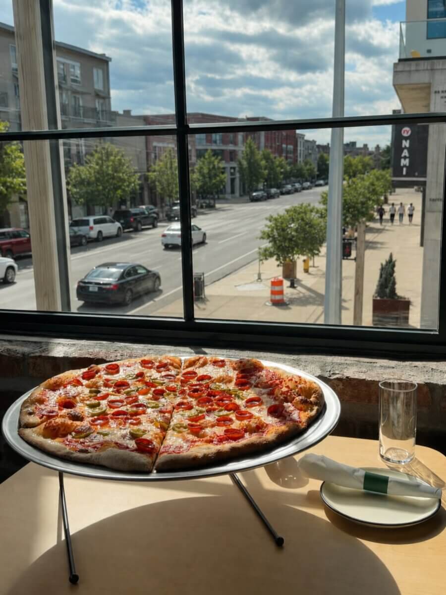 25+ Louisville Restaurants Open on July 4th - 7 A pepperoni pizza sits on a stand by a window overlooking a city street; an empty plate, glass, and wrapped utensils are on the table in one of the Louisville restaurants open July 4th.