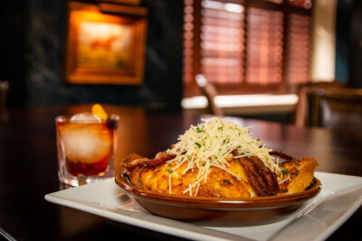 A plate of savory bread pudding topped with shredded cheese and herbs sits on a table beside a cocktail with ice and an orange twist—perfect for celebrating July 4th at open Louisville restaurants.