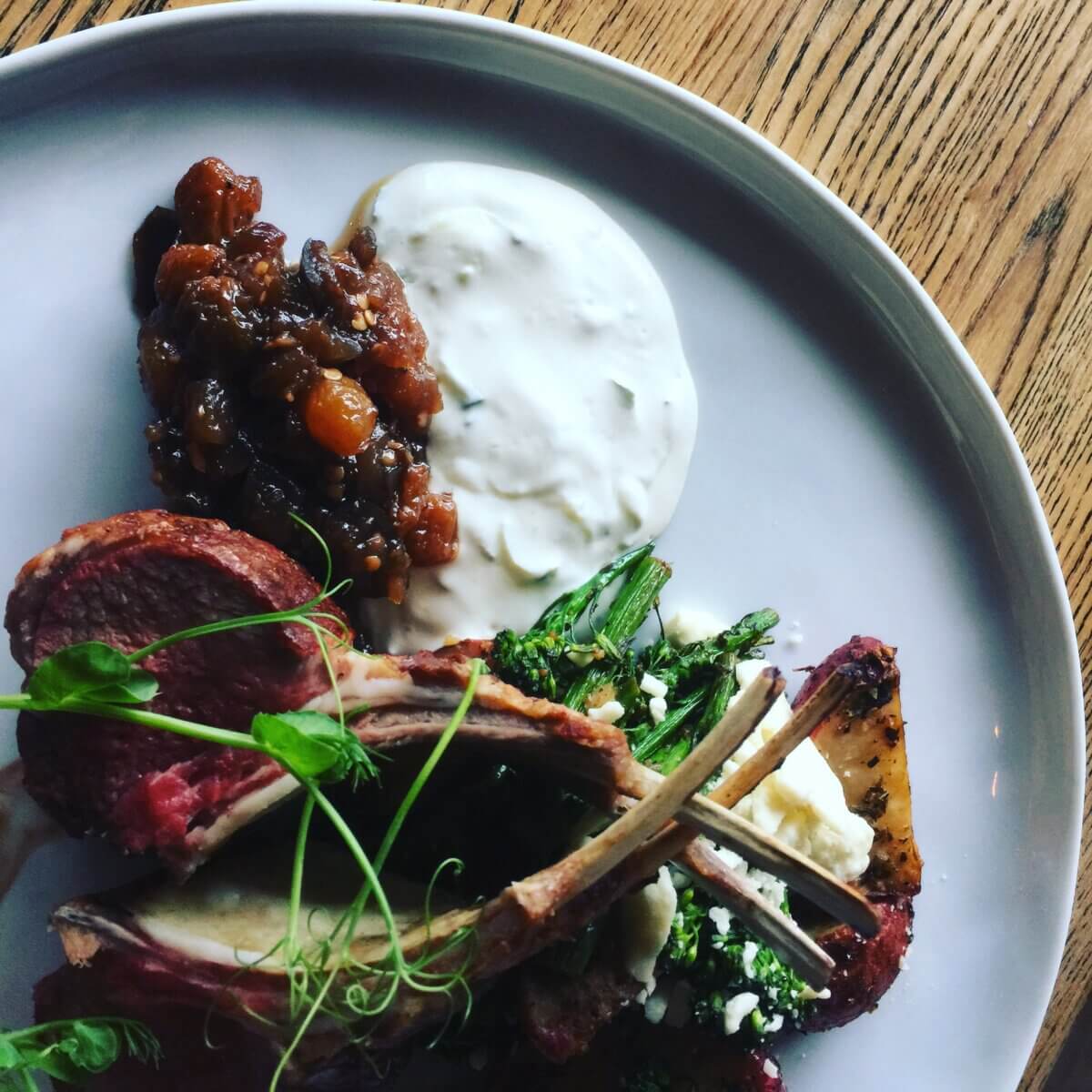 25+ Louisville Restaurants Open on July 4th - 2 Plate with rack of lamb, broccolini, a dollop of white sauce, a portion of chutney, and a garnish, served on a wooden table.