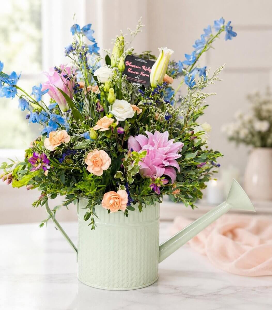 Your Guide to U-Pick Flower Farms Around Louisville - 2 A pastel green watering can filled with a mixed bouquet of pink, blue, white, and orange flowers sits on a marble countertop.