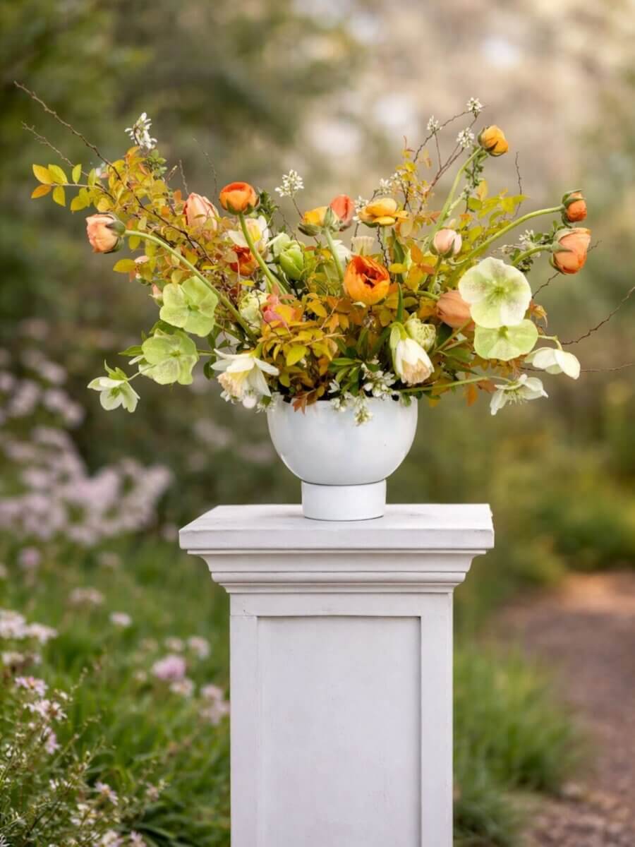 Your Guide to U-Pick Flower Farms Around Louisville - 4 A white vase with an arrangement of orange, yellow, and green flowers sits on a white pedestal outdoors, with blurred greenery in the background.