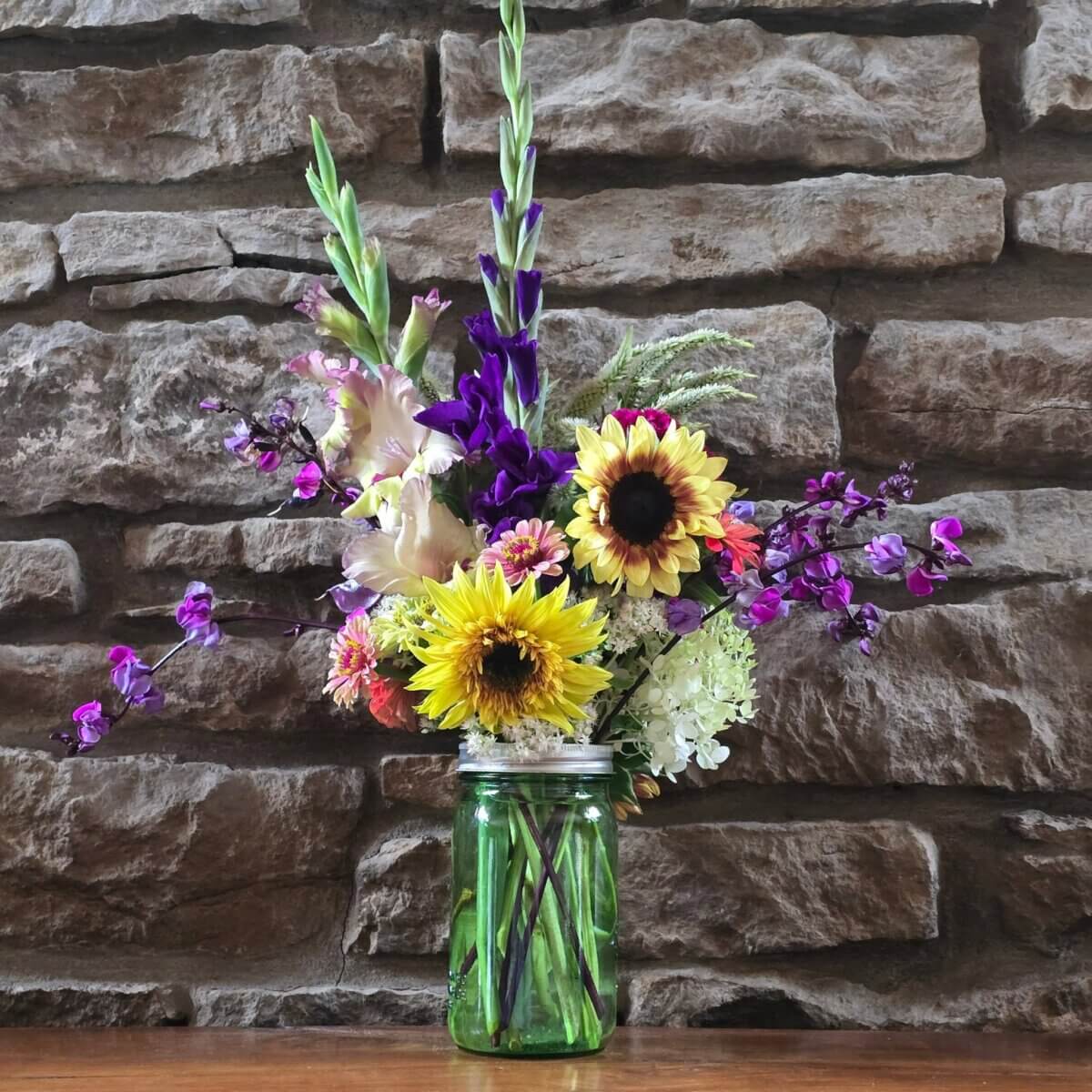 Your Guide to U-Pick Flower Farms Around Louisville - 6 A bouquet of colorful flowers, including sunflowers and purple blooms, arranged in a green mason jar, set on a wooden surface in front of a stone wall.