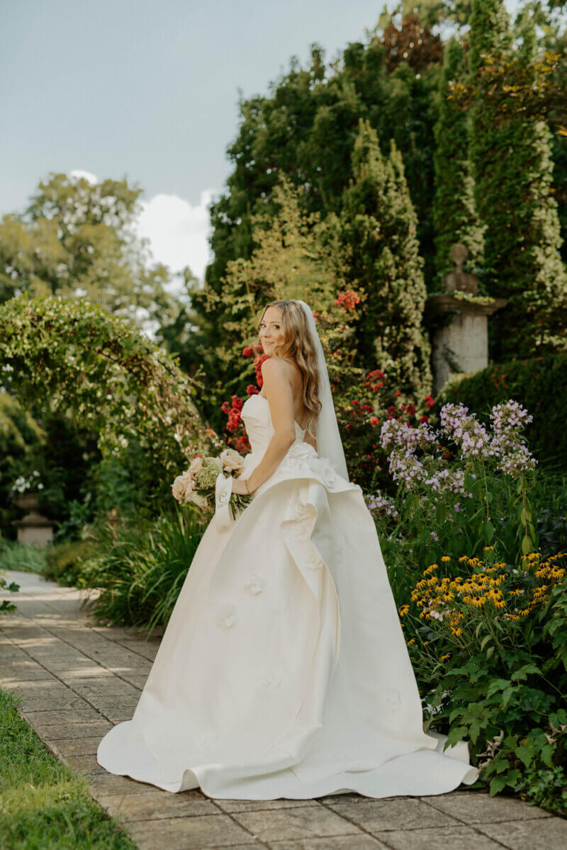 This Summer Wedding Was Straight Out of a Rom-Com - 3 A bride in a white gown holding a bouquet stands on a garden pathway at her Whitehall wedding, surrounded by green foliage and colorful flowers.