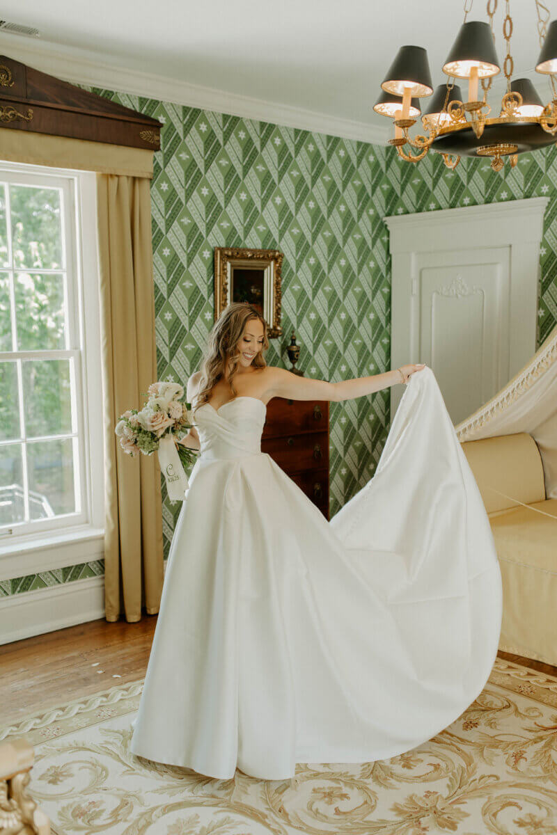This Summer Wedding Was Straight Out of a Rom-Com - 5 A bride in a strapless white gown holds a bouquet and lifts her dress in a green-patterned room with a large window, chandelier, and elegant furnishings—a perfect scene for a timeless Louisville wedding.