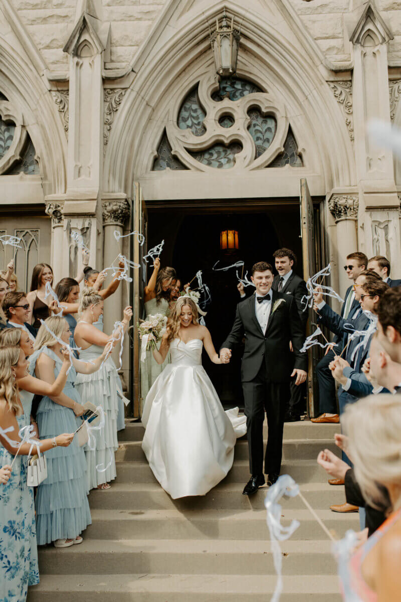 This Summer Wedding Was Straight Out of a Rom-Com - 7 A newlywed couple walks down church steps, holding hands, while guests on both sides celebrate and wave streamers—capturing the joy of their Louisville wedding love story.