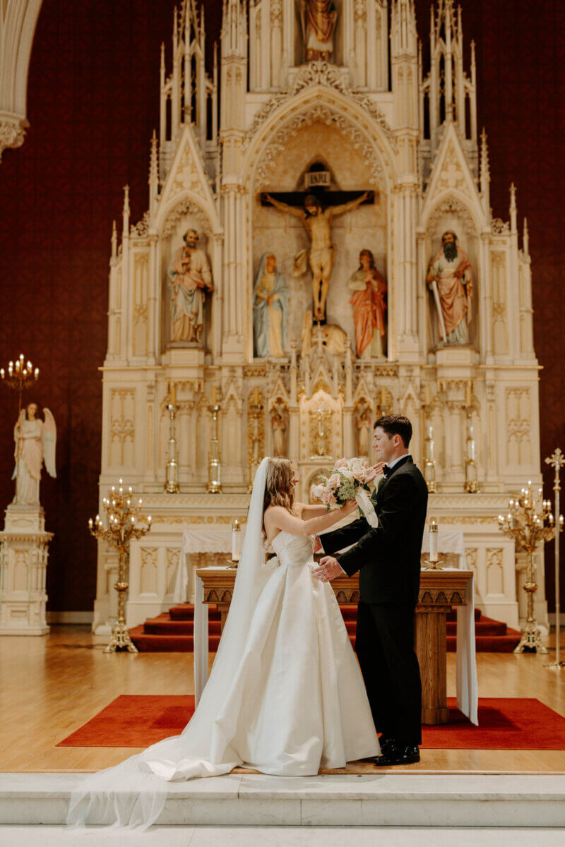 This Summer Wedding Was Straight Out of a Rom-Com - 6 A bride and groom stand facing each other at the altar of an ornate church, holding hands and surrounded by religious statues—a timeless Louisville wedding capturing their beautiful love story.