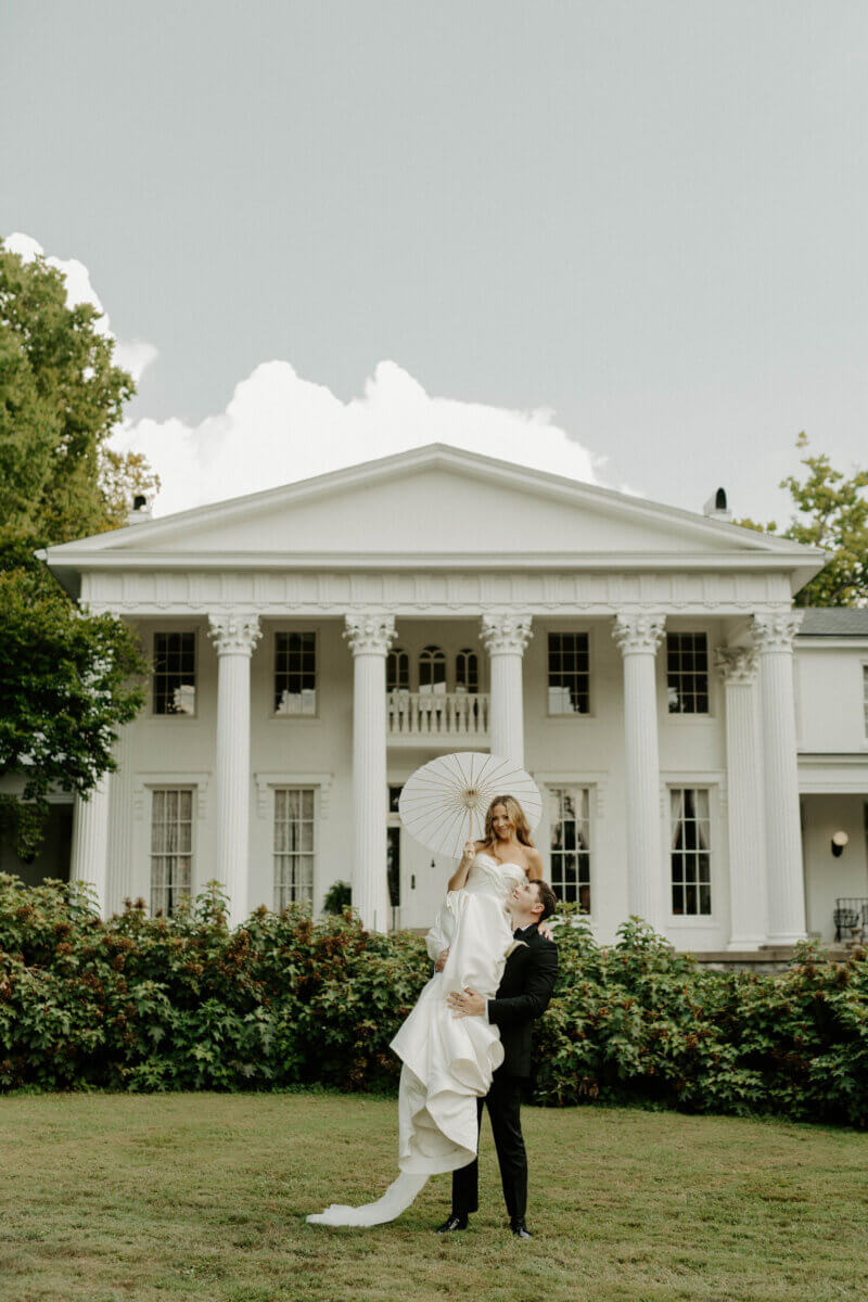 This Summer Wedding Was Straight Out of a Rom-Com - 4 A bride holding a white parasol is lifted by the groom in front of the grand Whitehall mansion, capturing a magical moment in their Louisville wedding love story on the lush, grassy lawn.