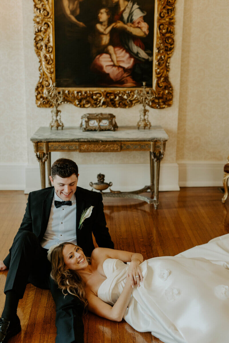 This Summer Wedding Was Straight Out of a Rom-Com - 2 A bride in a strapless white gown lies on the floor, smiling, next to a groom in a black tuxedo, capturing a beautiful Louisville wedding love story with a table and ornate framed painting in the background.