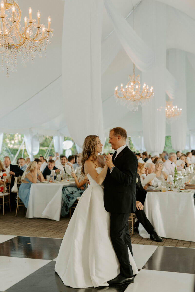 This Summer Wedding Was Straight Out of a Rom-Com - 13 A bride and groom share their first dance in a white tented venue at a Whitehall wedding, surrounded by seated guests and chandeliers.