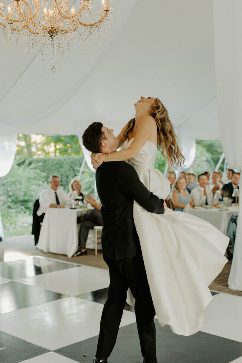 This Summer Wedding Was Straight Out of a Rom-Com - 12 A groom in a black suit lifts his bride in a white dress as they dance on a black-and-white floor, while guests smile under a chandelier and white tent—capturing the magic of their Louisville wedding and unforgettable love story.