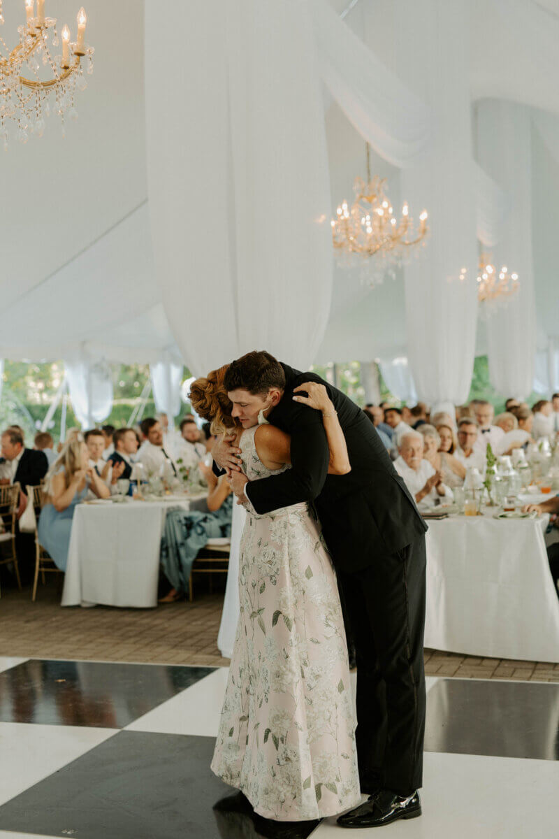 This Summer Wedding Was Straight Out of a Rom-Com - 14 A man in a black suit hugs a woman in a floral dress on the dance floor at a Louisville wedding, with guests seated at tables in the background, capturing a special moment in their love story.