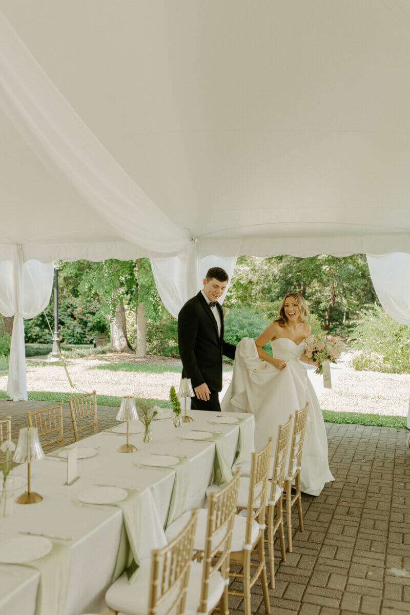 This Summer Wedding Was Straight Out of a Rom-Com - 8 A bride and groom stand beside a set dining table under a white tent outdoors at their Whitehall wedding, with greenery visible in the background.