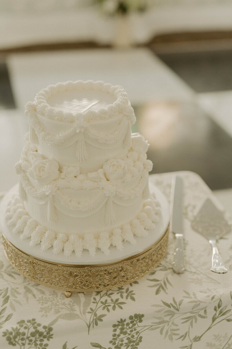 This Summer Wedding Was Straight Out of a Rom-Com - 11 Two-tier white frosted cake with ornate piping and floral decorations on a gold stand, placed on a patterned tablecloth with a cake knife and server—perfect for a Louisville wedding and celebrating your unique love story.