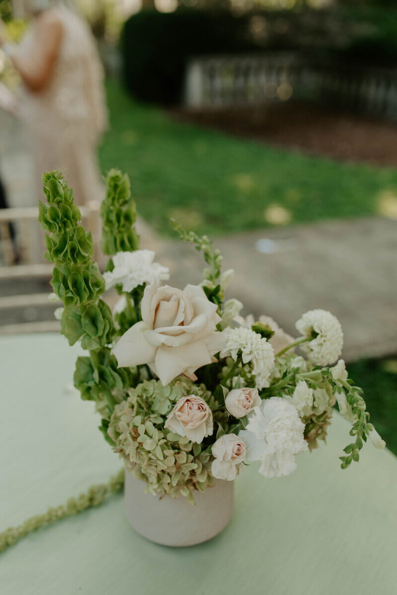 This Summer Wedding Was Straight Out of a Rom-Com - 9 A ceramic vase filled with white and pale pink roses, carnations, and green foliage sits on a light-colored table outdoors, perfect for a Whitehall wedding or any love story set in an elegant Louisville wedding garden.