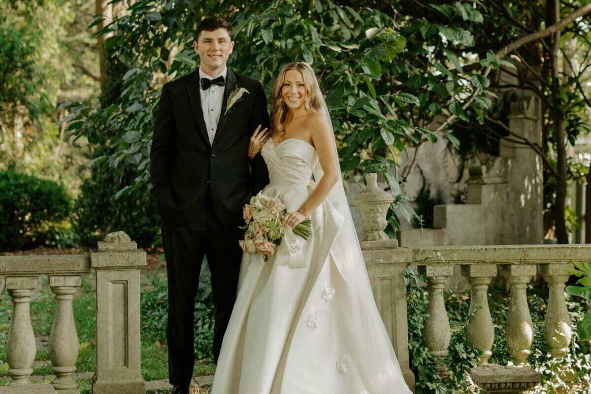 A bride in a strapless white gown holding a bouquet stands beside a groom in a black tuxedo, posing outdoors by a stone railing with greenery—a beautiful moment from their Whitehall wedding and timeless love story.
