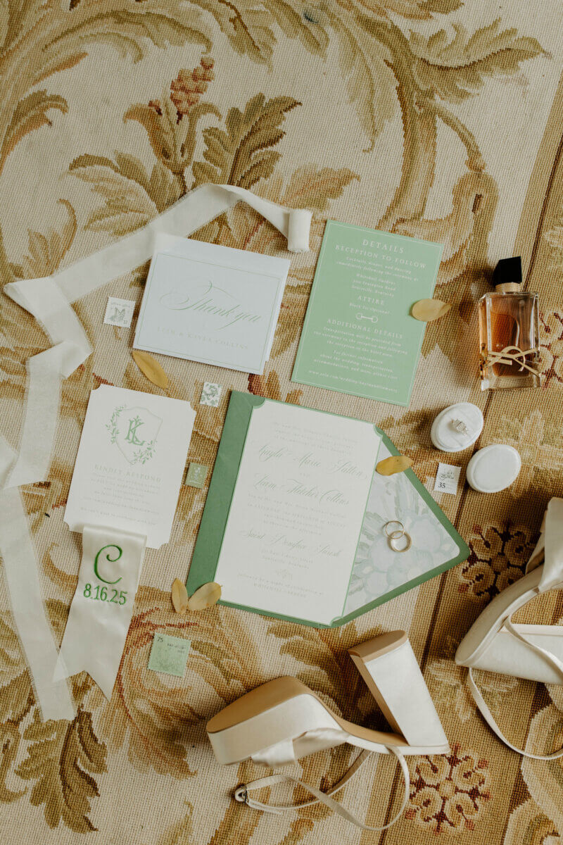 This Summer Wedding Was Straight Out of a Rom-Com - 10 Flat lay of a Whitehall wedding invitation suite, green and white cards, perfume bottle, wedding rings, ribbon, white high heels, and small decorative items on a patterned rug—capturing the start of a beautiful love story.