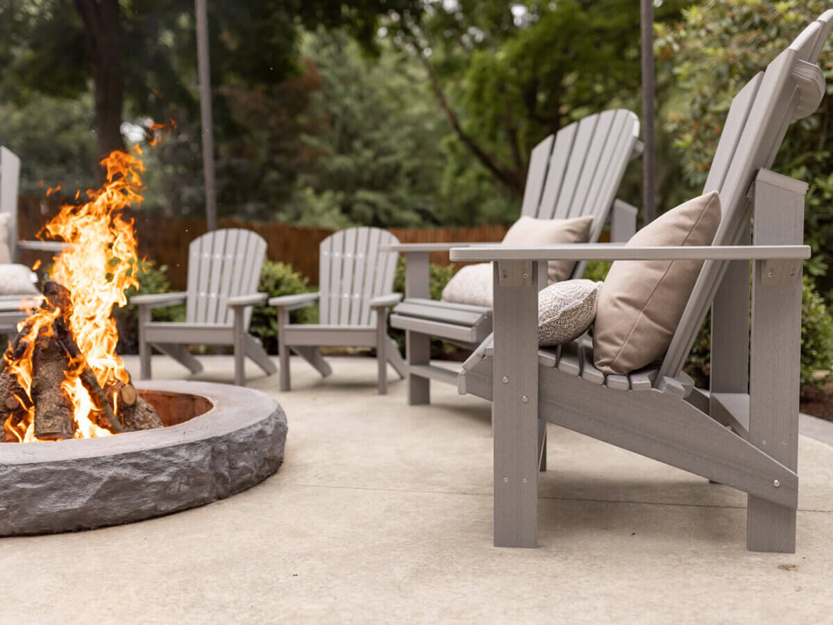 How to Refresh Your Porch in One Weekend, According to the Experts - 7 Gray Adirondack chairs with beige cushions surround a stone fire pit on a concrete patio, perfect for a porch makeover. Trees and greenery in the background create a relaxing setting to refresh your porch.