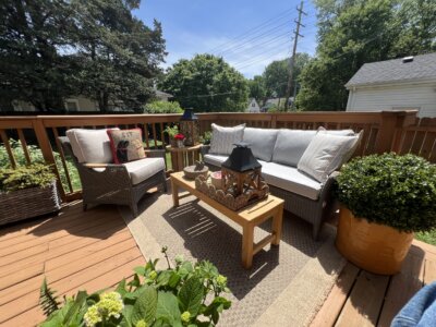 Outdoor patio setup with a cushioned sofa, armchair, coffee table, decorative pillows, potted plants, and lamps on a wooden deck under a sunny sky.