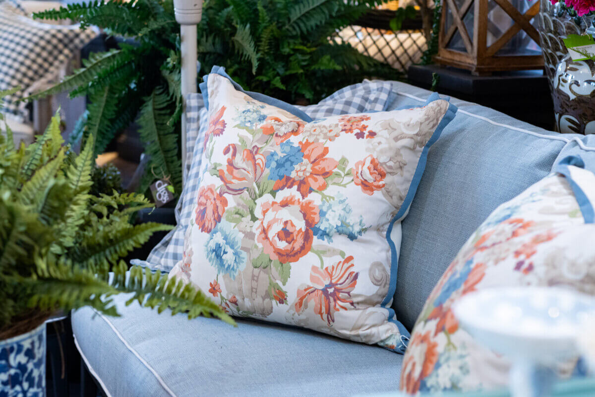 How to Refresh Your Porch in One Weekend, According to the Experts - 5 A floral-patterned pillow rests on a light blue couch, surrounded by green potted plants and decorative itemsβthe perfect touch for a weekend porch makeover inspired by Louisville experts.