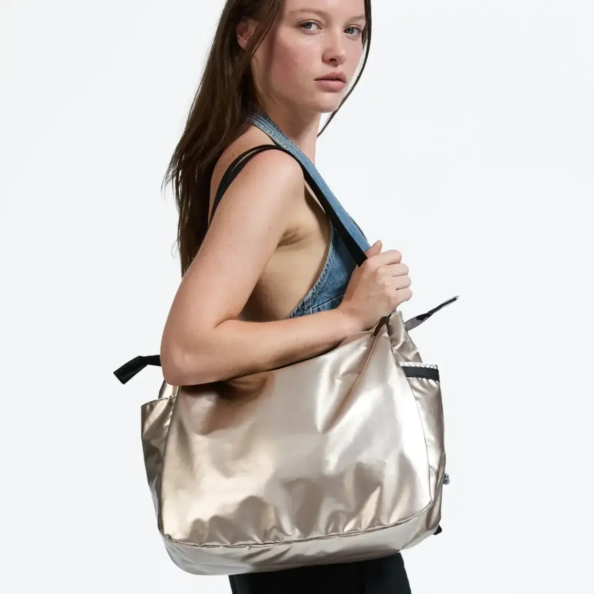 17 Luxurious Birmingham FINDS: Polished Summer Style, Artful Decor & Everyday Luxuries - 12 A woman with long brown hair, dressed in a sleeveless denim top, carries a luxurious, shiny metallic tote bag over her shoulder—perfect for summer outings in Birmingham—set against a plain background.