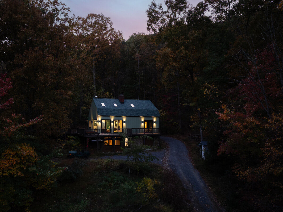 A two-story house with illuminated windows is surrounded by dense trees at dusk, with a gravel driveway leading to the entrance of this charming barn venue.