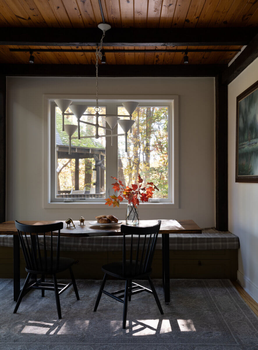 A wooden table with two black chairs is set by a window at The Marshall Barn, holding a vase with autumn leaves and a loaf of bread. Light filters in, highlighting a cozy, rustic dining nook.