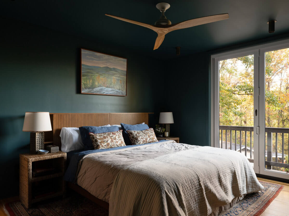 A modern bedroom boasts a large bed, two nightstands with lamps, a ceiling fan, and a landscape painting above the headboard, while glass doors overlook serene trees.